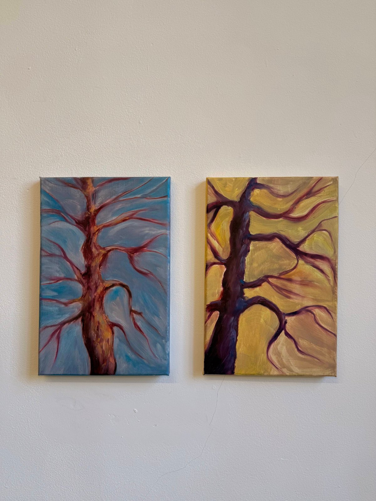 Two unframed canvases hanging side by side on an off-white wall: the left panel shows a bare tree in reddish-brown against a blue-grey sky, the right panel shows a dark plum tree against a warm gold background